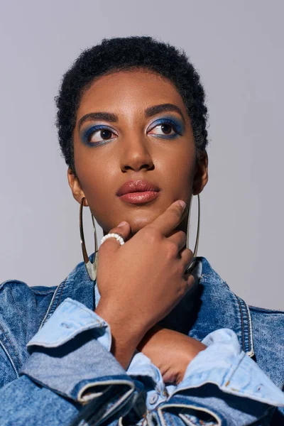 Portrait of trendy african american model with short hair and bold makeup wearing denim jacket and touching chin while looking away isolated on grey, denim fashion concept — Stock Photo