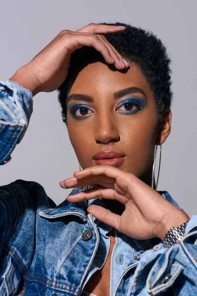 Portrait of modern african american woman with short hair and bold makeup touching face and looking at camera while posing in denim outfit isolated on grey, denim fashion concept — Stock Photo