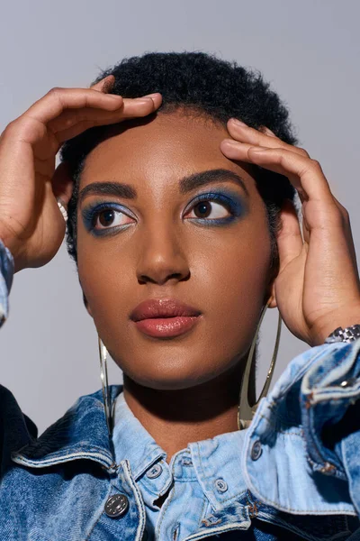 Portrait of stylish african american model with bold makeup and golden earrings touching short hair and looking away while posing in denim outfit isolated on grey, denim fashion concept — Stock Photo
