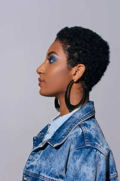Vista lateral de la mujer afroamericana moderna con pelo corto y maquillaje vívido que lleva pendientes de oro y chaqueta de mezclilla aislada en gris, concepto de moda denim - foto de stock