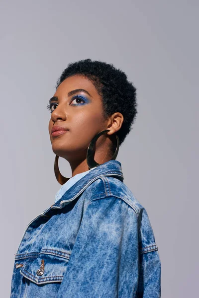 Low angle view of fashionable young african american woman with bold makeup posing in golden earrings and denim jacket while looking away isolated on grey, denim fashion concept — Stock Photo