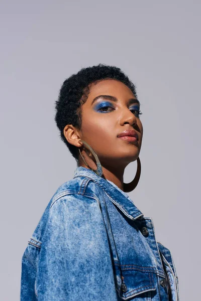 Low angle view of modern african american woman with vivid makeup and golden earrings looking at camera while posing in denim jacket isolated on grey, denim fashion concept — Stock Photo