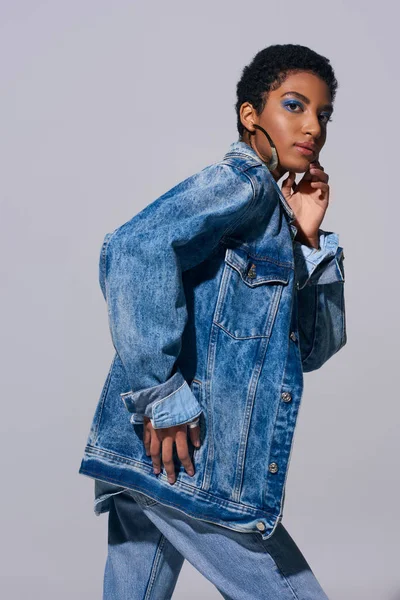 Modern african american model with bold makeup and short hair touching chin and looking at camera while posing in denim outfit isolated on grey, denim fashion concept — Stock Photo