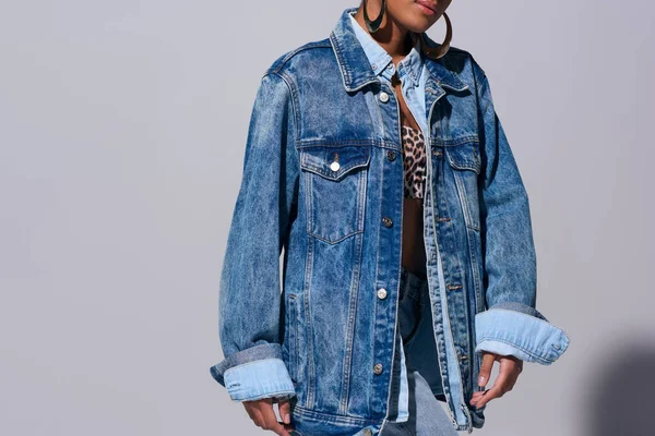 Розбитий вид модної молодої афро-американської жінки з золотими сережками поставлені зверху з тваринним друком і джинсовим піджаком на сірому фоні, денімська мода концепція — стокове фото