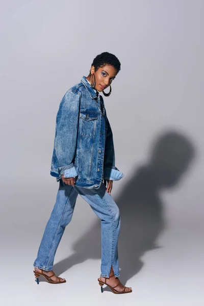 Piena lunghezza della giovane donna afroamericana alla moda con orecchini dorati guardando la fotocamera mentre posa in jeans e giacca di jeans su sfondo grigio, concetto di moda denim — Foto stock