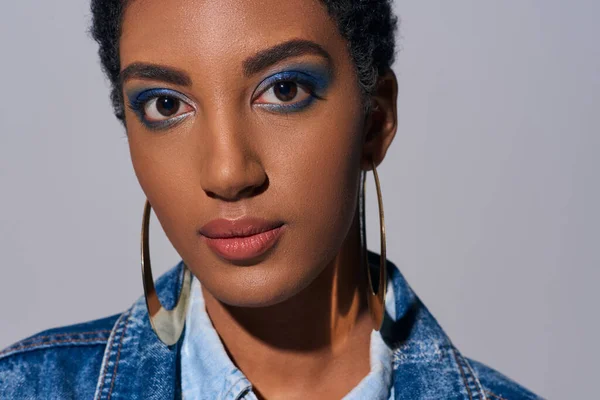Close up view of confident african american woman with vivid makeup and golden earrings looking at camera while wearing denim jacket isolated on grey, denim fashion concept — Stock Photo