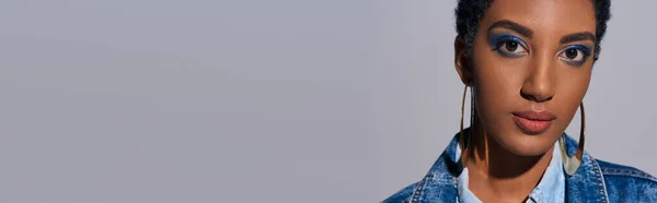 Ritratto di modello afroamericano corto dai capelli corti ed elegante con trucco audace e orecchini dorati che guardano la macchina fotografica mentre indossa giacca di denim isolata su grigio, concetto di moda denim, banner — Foto stock