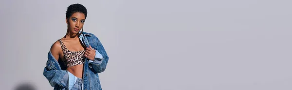 Femme afro-américaine moderne aux cheveux courts en haut avec imprimé animal et vêtements en denim regardant la caméra et debout sur fond gris, concept de mode en denim, bannière — Photo de stock