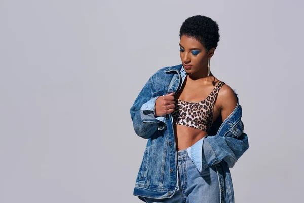 Fashionable african american woman with bold makeup wearing top with animal print and touching denim jacket while standing isolated on grey, denim fashion concept — Stock Photo
