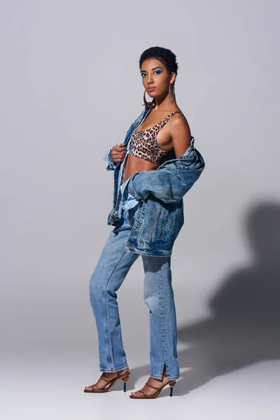 Piena lunghezza della donna afroamericana alla moda con trucco audace indossando top con stampa animalier, giacca in denim e jeans su sfondo grigio, concetto di moda denim — Foto stock