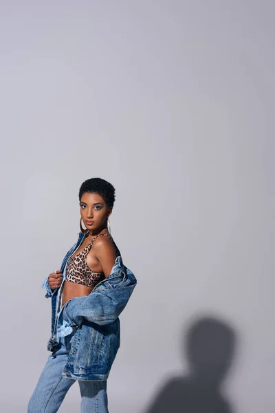Модная африканская американка с золотыми сережками и короткими волосами, позирующая сверху с изображением животных и джинсовой курткой на сером фоне, джинсовая концепция моды — стоковое фото