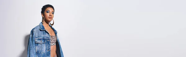 Donna afroamericana di tendenza con trucco vivido e orecchini dorati che indossa top con stampa animalier e giacca in denim su sfondo grigio con spazio copia, concetto di moda denim, banner — Foto stock