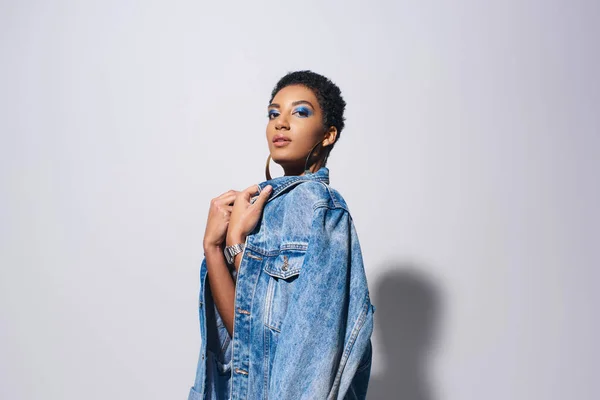 Confident african american model with short hair and golden earrings looking at camera while touching denim jacket and standing on grey background, denim fashion concept — Stock Photo