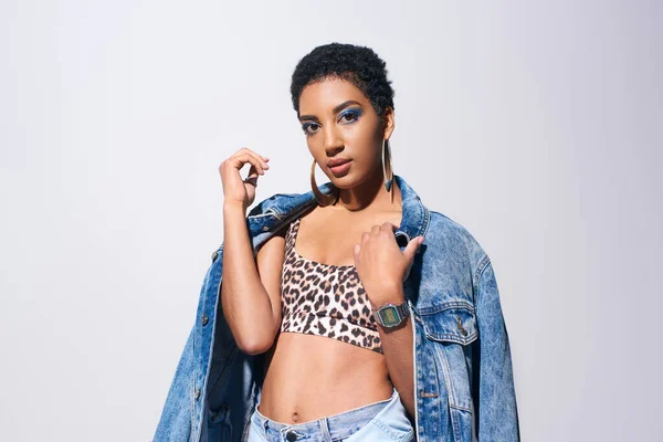 Trendy african american young woman with short hair and bold makeup wearing top with animal print and modern denim jacket while standing isolated on grey, denim fashion concept — Stock Photo