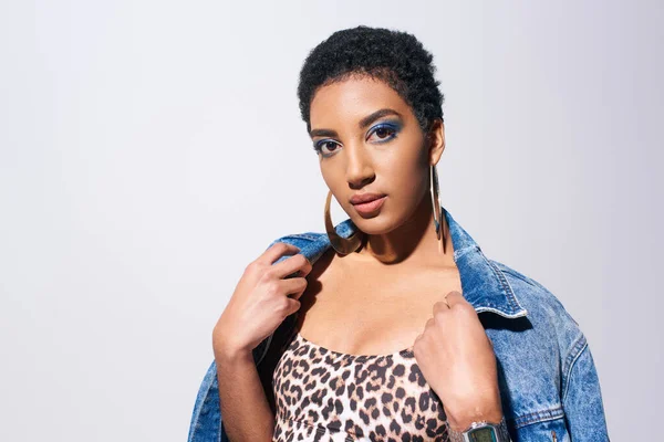 Young and short haired african american woman with blue eyeshadow and golden earrings posing in top with animal print and wearing denim jacket isolated on grey, denim fashion concept — Stock Photo