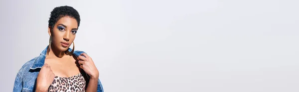 Mulher americana africana elegante com maquiagem vívida e brincos dourados vestindo jaqueta de ganga enquanto posando em cima com impressão animal isolada no conceito de moda cinza, ganga, banner — Fotografia de Stock
