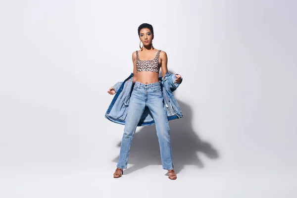 Full length of trendy and short haired african american model in top with leopard print and jeans posing with denim jacket while standing on grey background, denim fashion concept — Stock Photo