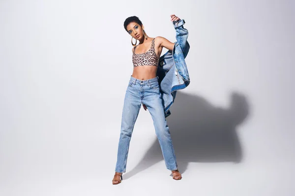 Стильная африканская модель с смелым макияжем и золотыми сережками, смотрящая в камеру, позируя в джинсовой куртке и джинсах на сером фоне, джинсовая концепция моды — стоковое фото