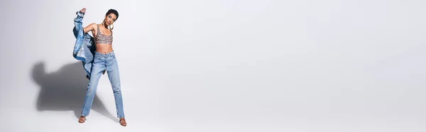 Modello afroamericano con capelli corti e orecchini dorati in posa in giacca di denim, top con stampa animalier e jeans su fondo grigio, concept moda denim, banner — Foto stock