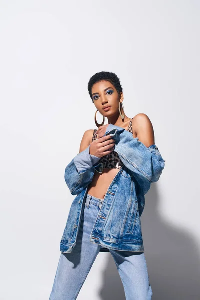 Trendy short haired african american model with golden earrings posing in denim jacket and jeans while looking at camera on grey background, denim fashion concept — Stock Photo