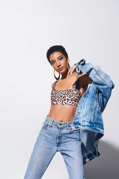 Confident african american woman with vivid makeup in top with animal print and jeans wearing denim jacket and posing on grey background, denim fashion concept — Stock Photo