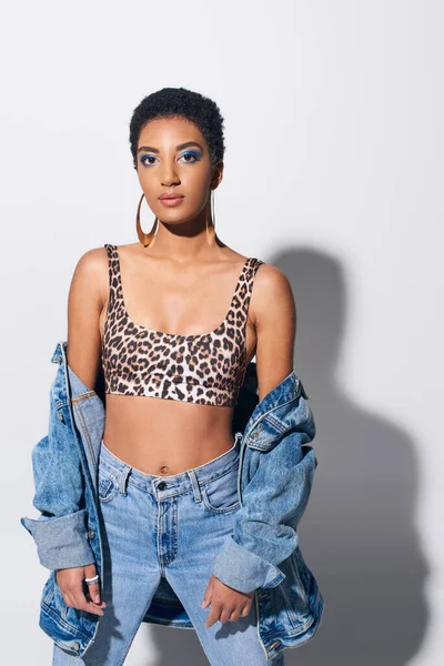 Trendy young african american woman with bold makeup and golden earrings looking at camera and posing in top with animal print and denim outfit on grey background, denim fashion concept — Stock Photo