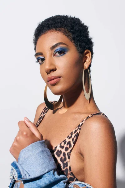 Portrait of short haired african american woman with bold makeup and golden earrings posing in top with animal print and denim jacket on grey background, denim fashion concept — Stock Photo