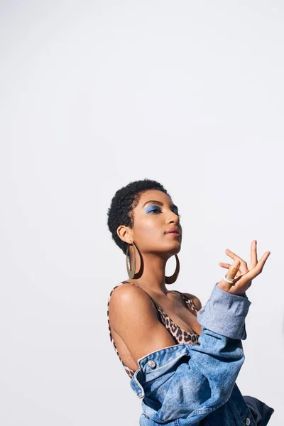 Young short haired african american woman with bold makeup posing in top with animal print and denim jacket while looking away isolated on grey, denim fashion concept — Stock Photo