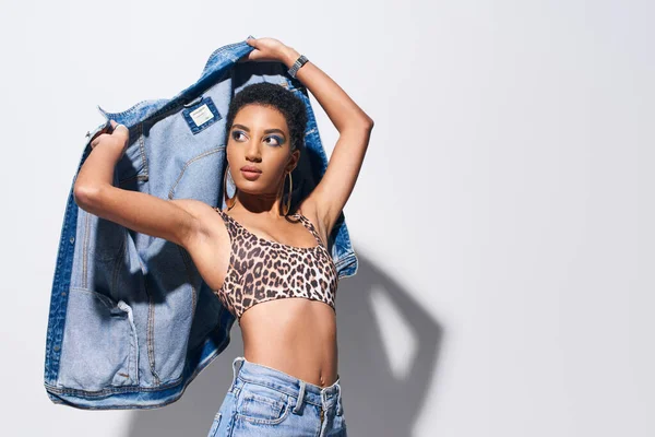 Jeune femme afro-américaine à la mode avec un maquillage audacieux et des boucles d'oreilles dorées tenant une veste en denim tout en posant en haut avec un imprimé animal et un jean sur fond gris, concept de mode en denim — Photo de stock