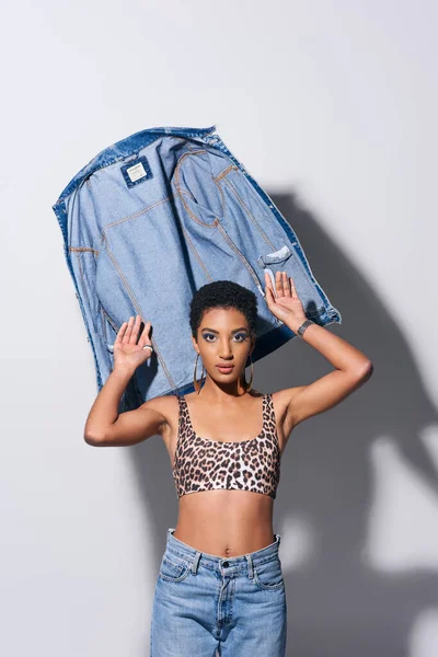 Тенді-афроамериканська модель з коротким волоссям зверху з малюнком леопарда і джинсами, які дивляться на камеру біля джинсової куртки на сірому тлі, денімська мода концепція — стокове фото