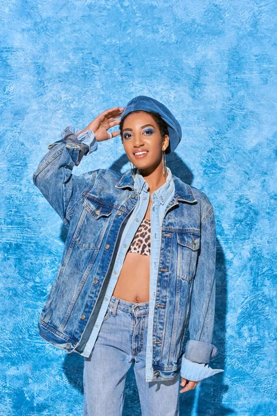 Cheerful and young african american woman with bold makeup touching beret while posing in denim jacket and jeans on blue textured background, stylish denim attire — Stock Photo