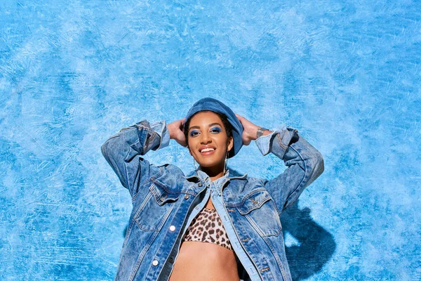 Positive and trendy young african american model in denim jacket and top with animal print touching beret and standing on blue textured background, stylish denim attire — Stock Photo