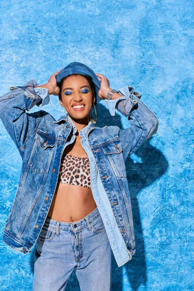 Trendy african american woman with closed eyes and bold makeup touching beret and posing in denim clothes and top on blue textured background, stylish denim attire — Stock Photo