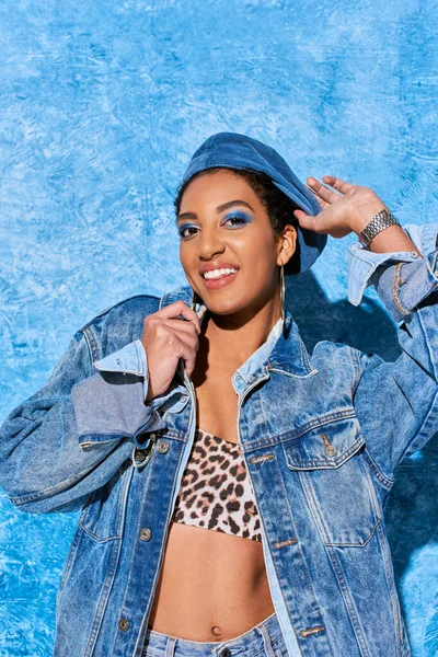 Joyful african american model in denim jacket and top with animal print touching beret and looking at camera on blue textured background, stylish denim attire — Stock Photo