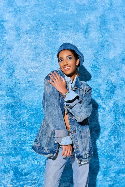 Jeune femme afro-américaine souriante avec un maquillage vif et un béret posant en jeans et veste en denim tout en se tenant debout sur un fond texturé bleu, tenue en denim élégante — Photo de stock