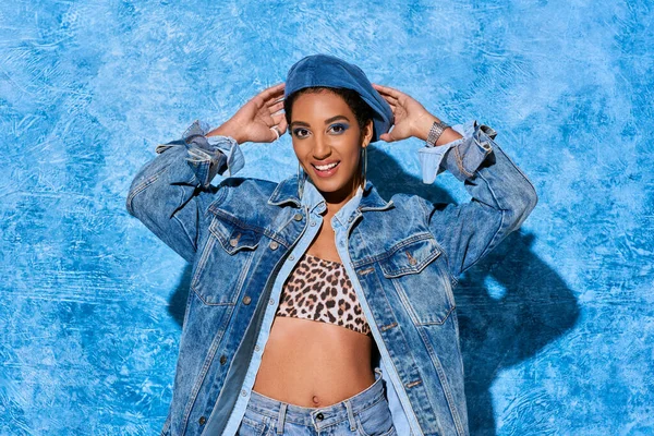 Впевнена і усміхаючись афро-американській жінці, яка торкається берету, одягаючись зверху з написом тварин і джинсовою курткою, стоячи на синьому текстурованому тлі, стильний денім. — стокове фото
