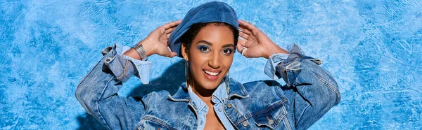 Retrato de modelo afroamericano sonriente y de pelo corto tocando la boina mientras posa en chaqueta de mezclilla y de pie sobre fondo azul texturizado, elegante atuendo de mezclilla, pancarta - foto de stock