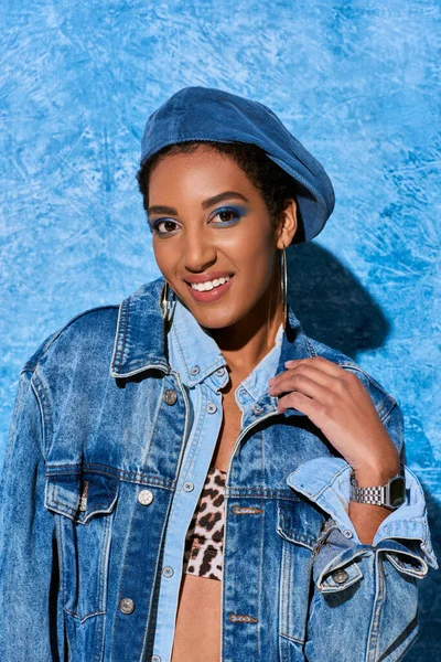 Cheerful african american model in beret, golden earrings and denim clothes looking at camera while standing near blue textured background, stylish denim attire — Stock Photo