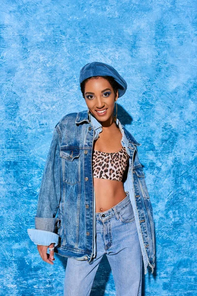 Positives afrikanisch-amerikanisches Model in Baskenmütze, Oberteil mit Leopardenmuster und Jeansjacke, die in die Kamera schaut und auf blauem strukturiertem Hintergrund steht, stylische Jeans-Kleidung — Stockfoto