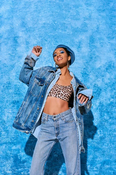 Low angle view of fashionable young african american model in beret posing in top and denim clothes while smiling at camera on blue textured background, stylish denim attire — Stock Photo
