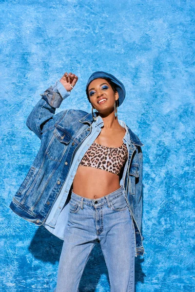 Positive african american model with bold makeup wearing beret, top with animal print and denim jacket while posing and standing near blue textured background, stylish denim attire — Stock Photo
