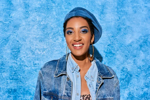 Retrato de mujer afroamericana joven y positiva con maquillaje audaz en boina y chaqueta de mezclilla mirando a la cámara sobre fondo azul texturizado, elegante atuendo de mezclilla - foto de stock