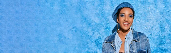 Modèle afro-américain joyeux avec fard à paupières audacieux et boucles d'oreilles dorées portant un béret et une veste en denim debout sur fond texturé bleu, tenue en denim élégante, bannière — Photo de stock