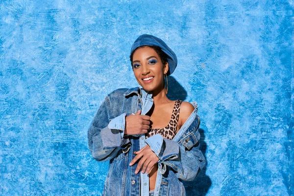 Mujer afroamericana de moda y sonriente con maquillaje audaz y boina posando en la parte superior con estampado de animales y chaqueta de mezclilla sobre fondo azul texturizado, elegante atuendo de mezclilla - foto de stock