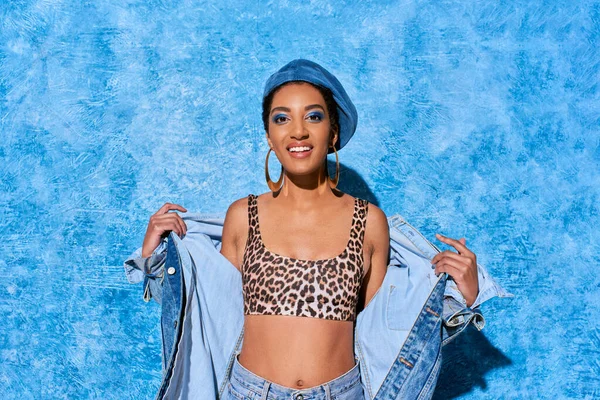Positive african american model with vivid makeup and beret posing in top with leopard print and denim outfit while standing on blue textured background, stylish denim attire — Stock Photo