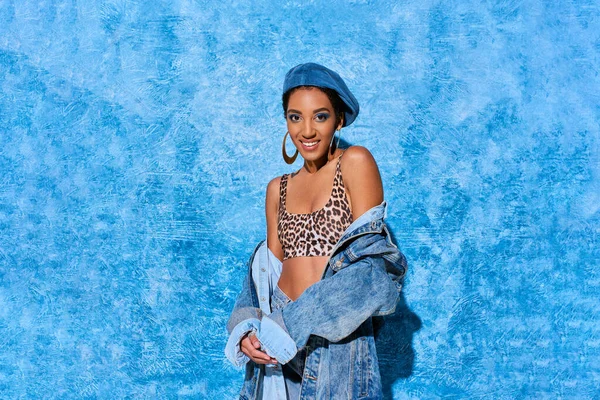 Fashionable african american woman in beret and top with animal print smiling at camera and posing in denim jacket on blue textured background, stylish denim attire — Stock Photo