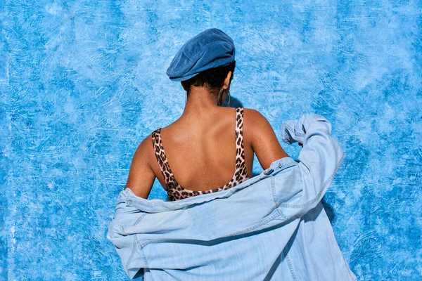 Vista posterior de la mujer afroamericana de pelo corto y de moda en boina, parte superior con estampado de animales y camisa de mezclilla de pie sobre fondo de textura azul, elegante atuendo de mezclilla - foto de stock