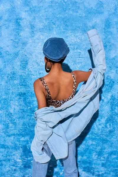 Visão traseira da mulher americana africana na moda em boina, top com impressão de leopardo e camisa de ganga em pé perto de fundo texturizado azul com sombra, traje de ganga elegante — Fotografia de Stock