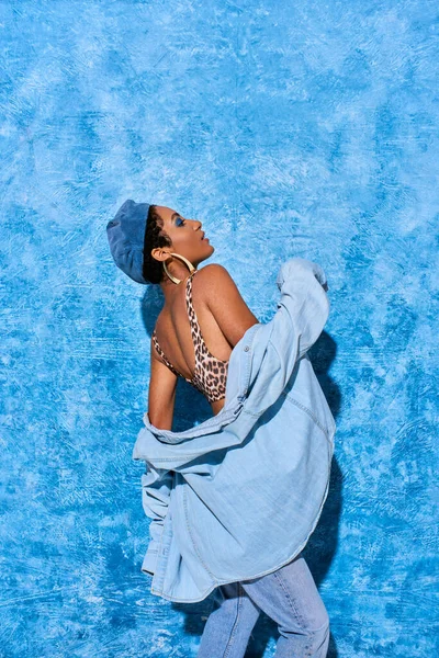 Vista laterale della donna afroamericana alla moda in berretto, top con stampa animalier e camicia in denim in posa e in piedi su sfondo blu strutturato, elegante abbigliamento in denim — Foto stock