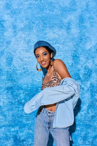 Модна афро-американська модель в золотих сережках, береті і джинсовій сорочці, посміхаючись на камеру, позуючи і стоячи на синьому текстурованому фоні, стильний джинсовий одяг — стокове фото
